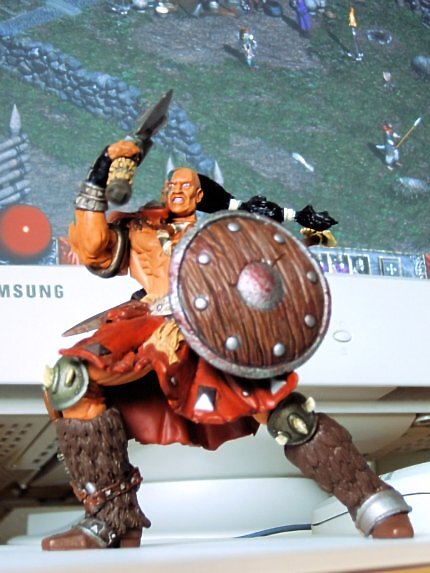 Eine Nahaufnahme einer detaillierten Diablo 2 Barbaren-Actionfigur aus Kunststoff. Der muskulöse Krieger mit Zopf, blauer Gesichtsbemalung, Fellstiefeln und rotem Lendenschurz posiert kampfbereit mit erhobenem Krummdolch und einem runden Holzschild. Im unscharfen Hintergrund steht ein heller Samsung-CRT-Röhrenmonitor, auf dessen Bildschirm das Gameplay von Diablo 2 im ersten Akt zu sehen ist, inklusive der roten Lebenskugel unten links.
