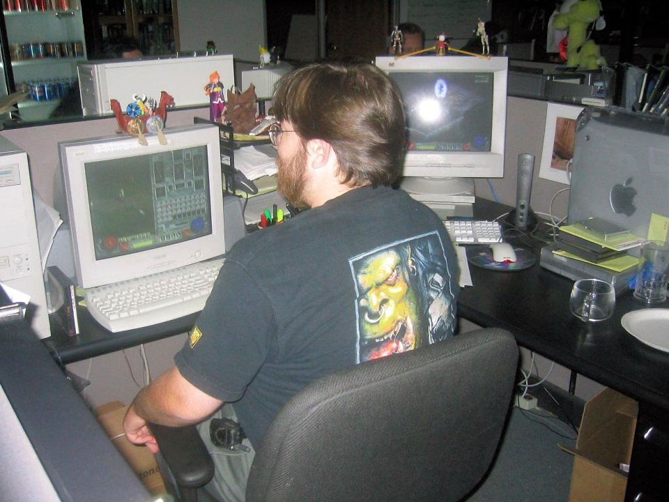 Ein historisches Foto aus einem Blizzard-Büro. Ein bärtiger Mitarbeiter mit Brille und einem schwarzen Warcraft-Orc-T-Shirt sitzt von hinten auf einem Bürostuhl vor zwei großen, weißen CRT-Röhrenmonitoren. Auf beiden Bildschirmen ist das Gameplay und das Interface von Diablo 2 geöffnet. Auf den Monitoren stehen kleine Spielfiguren, im Hintergrund sind typische Büro-Arbeitsplätze und Regale mit Getränkedosen zu sehen.